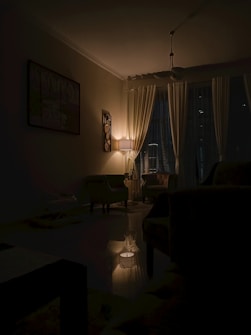 A dimly lit living room with a lamp casting a warm glow over the space. The room features curtains covering the windows, a painting on the wall, and comfortable chairs arranged around a central area. The ambiance is calm and intimate, with shadows enhancing the cozy atmosphere.