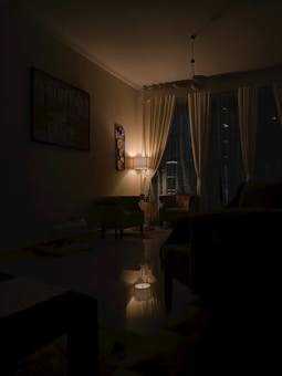 A dimly lit living room with a lamp casting a warm glow over the space. The room features curtains covering the windows, a painting on the wall, and comfortable chairs arranged around a central area. The ambiance is calm and intimate, with shadows enhancing the cozy atmosphere.
