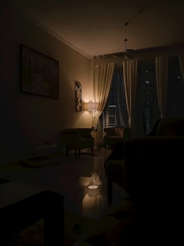 A dimly lit living room with a lamp casting a warm glow over the space. The room features curtains covering the windows, a painting on the wall, and comfortable chairs arranged around a central area. The ambiance is calm and intimate, with shadows enhancing the cozy atmosphere.