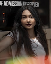 A person with long dark hair is in focus, standing indoors near a sign that partially reads 'OF ADMISSION RESERVED.' The person is wearing a white t-shirt with a small graphic print and a gold chain necklace. The image has a vintage effect with a timestamp and colorful light leaks at the bottom.