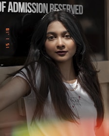 A person with long dark hair is in focus, standing indoors near a sign that partially reads 'OF ADMISSION RESERVED.' The person is wearing a white t-shirt with a small graphic print and a gold chain necklace. The image has a vintage effect with a timestamp and colorful light leaks at the bottom.