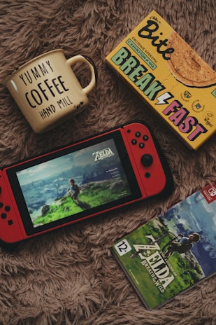 A cozy setup featuring a yellow mug with 'YUMMY COFFEE' written on it, a box of breakfast cookies, a red handheld gaming console displaying the game 'Zelda: Breath of the Wild', and the game case for the same. All items are placed on a soft, furry surface.
