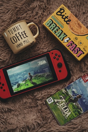 A cozy setup featuring a yellow mug with 'YUMMY COFFEE' written on it, a box of breakfast cookies, a red handheld gaming console displaying the game 'Zelda: Breath of the Wild', and the game case for the same. All items are placed on a soft, furry surface.