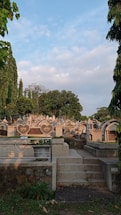 A cemetery features numerous gravestones with various shapes, surrounded by lush greenery and tall trees under a partly cloudy sky. A set of stone steps leads up to the area, and a metal railing with heart decoration encloses a planter.