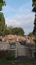 A cemetery features numerous gravestones with various shapes, surrounded by lush greenery and tall trees under a partly cloudy sky. A set of stone steps leads up to the area, and a metal railing with heart decoration encloses a planter.