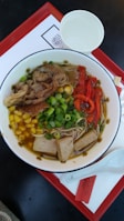 A bowl of ramen featuring sliced vegetables and meat with vibrant colors. Ingredients include sliced red bell peppers, chopped green onions, slices of tofu, corn kernels, and a portion of sliced meat, all in a rich broth accompanied by noodles. The dish is presented on a red rectangular tray alongside a white paper cup and a white ceramic spoon.