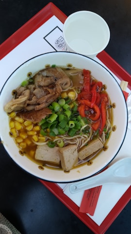 A bowl of ramen featuring sliced vegetables and meat with vibrant colors. Ingredients include sliced red bell peppers, chopped green onions, slices of tofu, corn kernels, and a portion of sliced meat, all in a rich broth accompanied by noodles. The dish is presented on a red rectangular tray alongside a white paper cup and a white ceramic spoon.