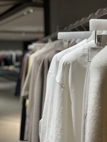A row of neatly hung clothes on hangers, featuring mainly neutral-toned garments such as whites and beiges. The background is softly blurred, suggesting a retail or wardrobe setting.