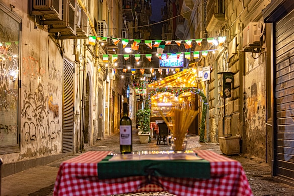 A cozy narrow street with warm yellow lighting and festive flags overhead. Graffiti decorates the walls. A checkered tablecloth with a bottle of wine and spaghetti is in the foreground, suggesting an inviting dining setup. Strings of lights and a welcoming restaurant sign create a charming atmosphere in the background.