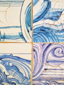 A set of ceramic tiles featuring artistic, abstract wave patterns in blue and white tones. Each tile displays a different style of swirling lines and curves, with visible cracks and a weathered look.