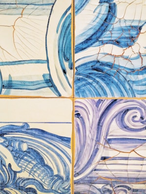 A set of ceramic tiles featuring artistic, abstract wave patterns in blue and white tones. Each tile displays a different style of swirling lines and curves, with visible cracks and a weathered look.