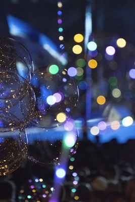 A collection of bubbles reflecting various colorful lights creates a mesmerizing visual display. Out-of-focus colorful circles of light form a bokeh effect in the background, enhancing the abstract and dreamy atmosphere. The image has a dark backdrop, making the bright bubbles and lights stand out prominently.