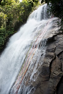 A waterfall cascades down a rocky cliff surrounded by lush green foliage. Colorful ropes are draped down the waterfall, possibly indicating a climbing or rappelling route.