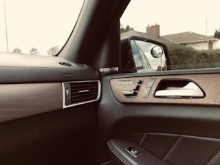 Interior of a car showing a side door panel with controls, a side mirror reflecting the exterior view of a house and greenery, and an air vent. The design includes wood and leather elements.