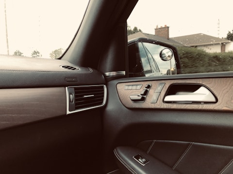 Interior of a car showing a side door panel with controls, a side mirror reflecting the exterior view of a house and greenery, and an air vent. The design includes wood and leather elements.