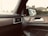Interior of a car showing a side door panel with controls, a side mirror reflecting the exterior view of a house and greenery, and an air vent. The design includes wood and leather elements.