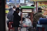 A street vendor stall is serving customers on a busy city sidewalk. The stall is labeled 'Sekoteng' and is equipped with a metal pot and various containers. Several people are gathered around the stall, some sitting and others standing, with face masks visible on some individuals. A colorful menu poster is displayed next to the stall.