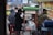 A street vendor stall is serving customers on a busy city sidewalk. The stall is labeled 'Sekoteng' and is equipped with a metal pot and various containers. Several people are gathered around the stall, some sitting and others standing, with face masks visible on some individuals. A colorful menu poster is displayed next to the stall.