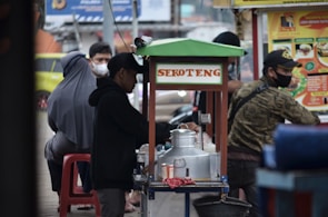 A street vendor stall is serving customers on a busy city sidewalk. The stall is labeled 'Sekoteng' and is equipped with a metal pot and various containers. Several people are gathered around the stall, some sitting and others standing, with face masks visible on some individuals. A colorful menu poster is displayed next to the stall.