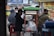 A street vendor stall is serving customers on a busy city sidewalk. The stall is labeled 'Sekoteng' and is equipped with a metal pot and various containers. Several people are gathered around the stall, some sitting and others standing, with face masks visible on some individuals. A colorful menu poster is displayed next to the stall.