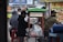 A street vendor stall is serving customers on a busy city sidewalk. The stall is labeled 'Sekoteng' and is equipped with a metal pot and various containers. Several people are gathered around the stall, some sitting and others standing, with face masks visible on some individuals. A colorful menu poster is displayed next to the stall.