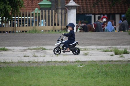 A child wearing a dark outfit and hat is riding a small toy motorcycle on a paved area, surrounded by grass. In the background, there are additional people, some in colorful attire, near a fence and a building with a tiled roof.