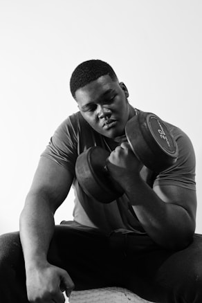A person intensely focuses on lifting a large dumbbell, sitting in a relaxed posture, wearing a casual t-shirt and pants. The image is in black and white, highlighting strong muscle definition and a determined expression.