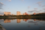 A serene lake reflects the image of several apartment buildings in the distance, surrounded by lush green trees. The sky is partly cloudy with warm lighting from the setting or rising sun.