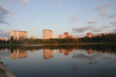 A serene lake reflects the image of several apartment buildings in the distance, surrounded by lush green trees. The sky is partly cloudy with warm lighting from the setting or rising sun.