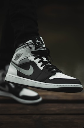 A close-up of a black and white sneaker with a distinct logo on the side, showcased outdoors on a textured wooden surface. The shoe features black laces and contrasting light and dark panels, contributing to its stylish design.