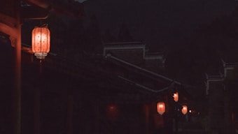 A night scene showcasing traditional Chinese lanterns softly glowing with a warm orange light, hanging along the edges of dark, silhouetted rooftops. The dimly lit setting adds a mysterious and serene ambiance.