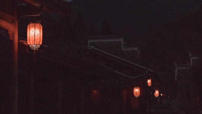 A night scene showcasing traditional Chinese lanterns softly glowing with a warm orange light, hanging along the edges of dark, silhouetted rooftops. The dimly lit setting adds a mysterious and serene ambiance.
