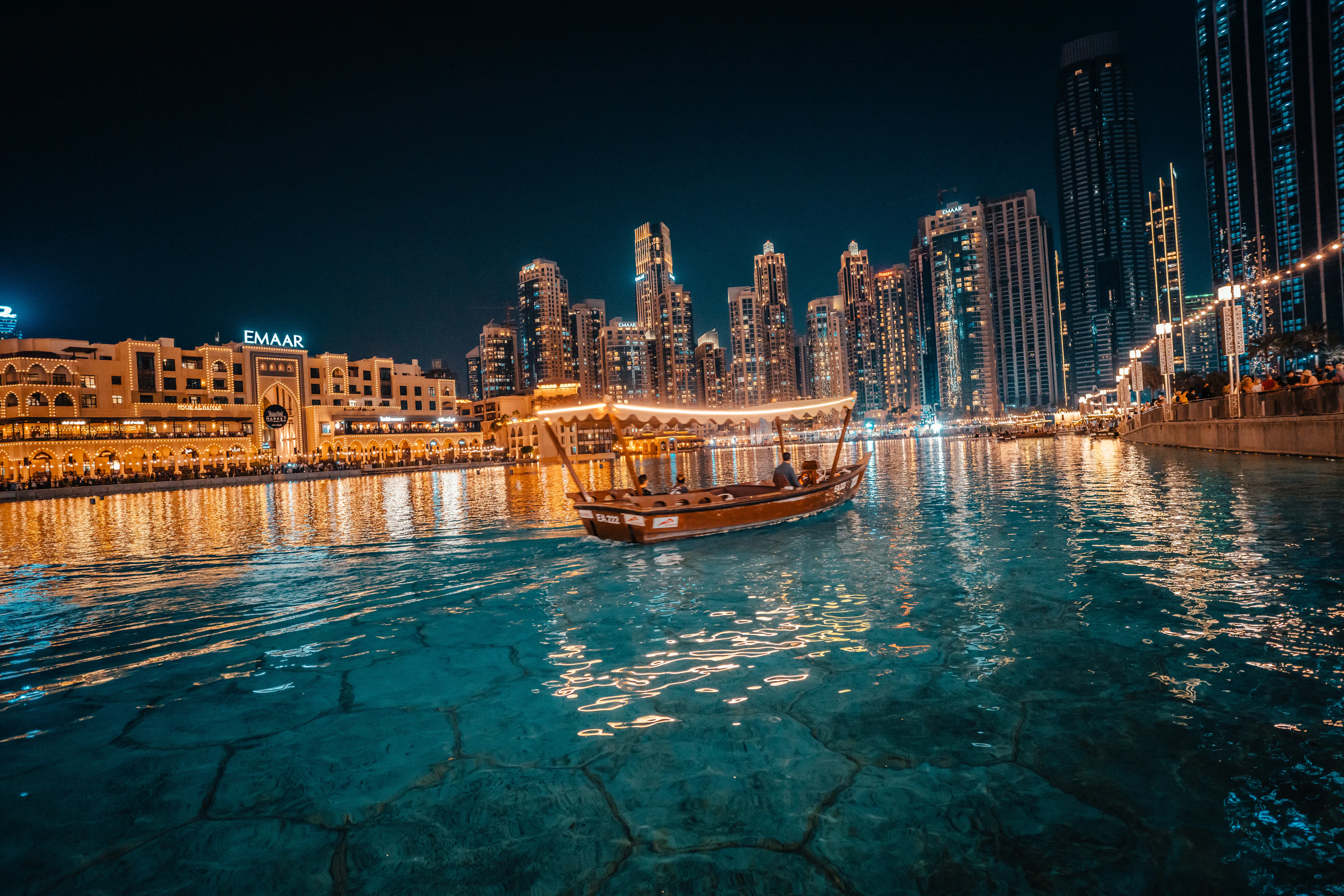 A wooden abra crossing the Burj Lake at night in Downtown Dubai, the towers of the promenade reflected in the water around it