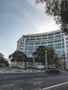 A modern multi-story glass office building with a curved facade is surrounded by trees. The street in front features a few moving vehicles and a paved pedestrian area. Illuminated letters spelling out a seasonal greeting are prominently displayed. The sky is clear with minimal cloud coverage, and some trees frame the top of the scene.