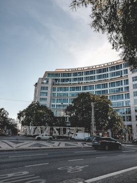 A modern multi-story glass office building with a curved facade is surrounded by trees. The street in front features a few moving vehicles and a paved pedestrian area. Illuminated letters spelling out a seasonal greeting are prominently displayed. The sky is clear with minimal cloud coverage, and some trees frame the top of the scene.