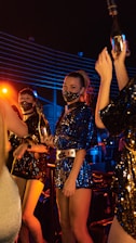 A group of people in a dimly lit party atmosphere wear sparkling sequin outfits and decorative face masks while holding bottles. The lighting is colorful, with blue and orange tones creating a vibrant and energetic environment.