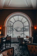 A cozy, elegantly furnished interior with leather chairs and a patterned carpet is visible. A large, arched window dominates the scene, providing a breathtaking view of snow-covered mountains and pine trees. Soft lighting from wall sconces adds a warm ambiance to the room.