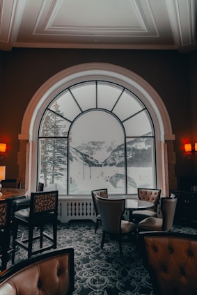 A cozy, elegantly furnished interior with leather chairs and a patterned carpet is visible. A large, arched window dominates the scene, providing a breathtaking view of snow-covered mountains and pine trees. Soft lighting from wall sconces adds a warm ambiance to the room.