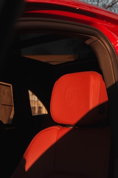 A close-up view of a luxurious car interior featuring a red leather seat, part of a headrest prominently visible. The headrest displays an embossed logo. Sunlight casts shadows across the seat, adding contrast. The car door frame is visible, coated in a bright red paint, and the background shows a hint of buildings softened by the light.