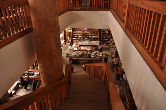 A spacious interior with wooden railings and a central staircase leads down to a retail area filled with various items such as kitchenware and pottery. Warm lighting and natural wood tones create a cozy ambiance.