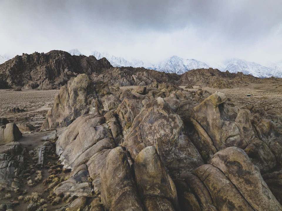 The image depicts a rocky landscape with a large boulder formation on the left side, surrounded by smaller rocks and boulders. The mountains in the background are covered in snow, creating a stark contrast against the sky. The overcast weather adds to the dramatic effect of the scene.
