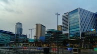 A modern urban area features several high-rise buildings characterized by glass facades. Notable structures have distinct designs, with patterns on the glass exterior or unique shapes. In the background, construction cranes are visible, indicating ongoing development. The environment appears to be a media complex as indicated by various logos on some of the buildings.