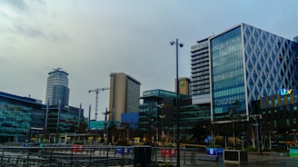 A modern urban area features several high-rise buildings characterized by glass facades. Notable structures have distinct designs, with patterns on the glass exterior or unique shapes. In the background, construction cranes are visible, indicating ongoing development. The environment appears to be a media complex as indicated by various logos on some of the buildings.
