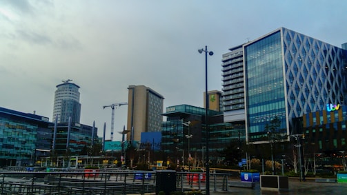 A modern urban area features several high-rise buildings characterized by glass facades. Notable structures have distinct designs, with patterns on the glass exterior or unique shapes. In the background, construction cranes are visible, indicating ongoing development. The environment appears to be a media complex as indicated by various logos on some of the buildings.
