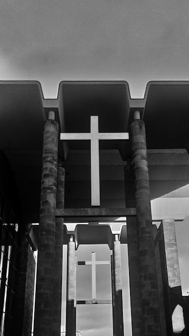 A black and white image of a modern architectural structure featuring a prominent cross. The structure includes tall stone columns supporting a large cross, with a backdrop of a cloudy sky. The design emphasizes symmetry and verticality.