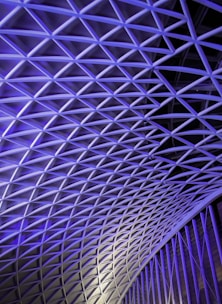 A complex geometric pattern of interlocking steel beams creates a lattice-like structure. The lighting casts a blue and purple hue across the surface, accentuating the modern architectural design.