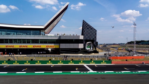 A modern racetrack setting with a distinctive building featuring a bold geometric design, including a large glass facade and angular roof structures. The track is wide and marked for racing, with advertisements like DHL and Rolex prominently displayed. A section of the track circuit is visible, set under a sky dotted with fluffy clouds. Spectators are gathered, adding to the dynamic and lively atmosphere.