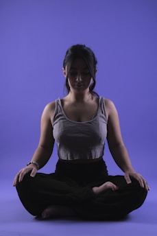 A person is sitting in a meditative pose with legs crossed on a smooth purple background. Their eyes appear closed, and they seem relaxed and focused. The lighting highlights the contours of their face and upper body, casting soft shadows.