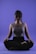 A person is sitting in a meditative pose with legs crossed on a smooth purple background. Their eyes appear closed, and they seem relaxed and focused. The lighting highlights the contours of their face and upper body, casting soft shadows.