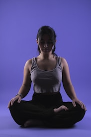 A person is sitting in a meditative pose with legs crossed on a smooth purple background. Their eyes appear closed, and they seem relaxed and focused. The lighting highlights the contours of their face and upper body, casting soft shadows.